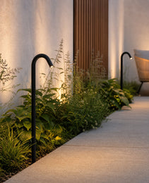 Black matte outdoor path light with curved neck casting warm LED glow on garden foliage