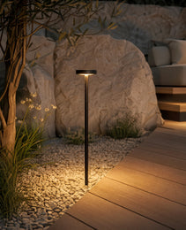 Black outdoor bollard light casting ambient warm white glow on gravel patio and stone boulders for residential landscape