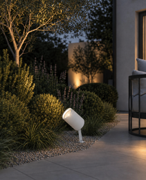 White matte outdoor spike lamp with angled head illuminating garden shrubs at dusk